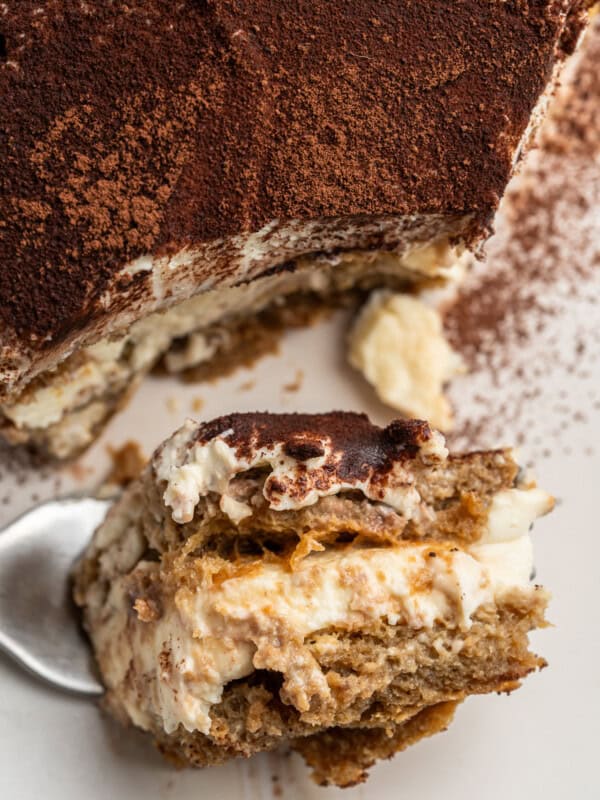 Close-up of a creamy tiramisu dessert on a white plate. A portion has been scooped out onto a spoon, revealing layers of espresso-soaked ladyfingers and mascarpone filling, dusted with cocoa powder.
