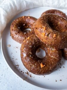 A close-up of a white plate with several glazed Pumpkin Spice Donuts, topped with orange zest and sprinkled with cinnamon. A light-colored cloth is partially visible in the background.
