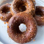 Four Pumpkin Spice Donuts with a brown, slightly glossy surface are stacked on a white plate. Small orange flecks peek through the donuts, and a white cloth is partially visible in the background.