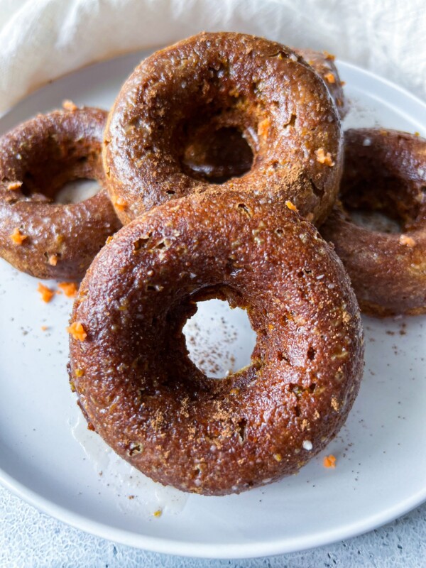 Four Pumpkin Spice Donuts with a brown, slightly glossy surface are stacked on a white plate. Small orange flecks peek through the donuts, and a white cloth is partially visible in the background.