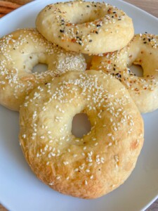 Four homemade bagels with golden crusts sit on a white plate. Made using an Easy Bagel Recipe, two are topped with sesame seeds and two with an everything seasoning blend.