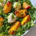 A Peach Burrata Salad featuring fresh arugula, grilled peach slices, creamy cheese dollops, and a garnish of herbs and edible flowers.