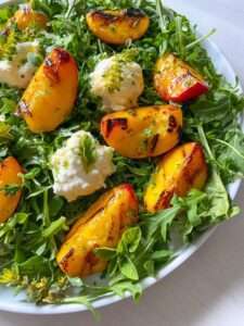 A Peach Burrata Salad featuring fresh arugula, grilled peach slices, creamy cheese dollops, and a garnish of herbs and edible flowers.