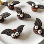 Halloween Chocolate Bats made from chocolate cookies with Oreo wings and candy eyes are arranged on a white plate. These whimsical treats look like playful bats, making them perfect for any Halloween celebration.