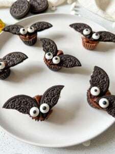 Halloween Chocolate Bats made from chocolate cookies with Oreo wings and candy eyes are arranged on a white plate. These whimsical treats look like playful bats, making them perfect for any Halloween celebration.