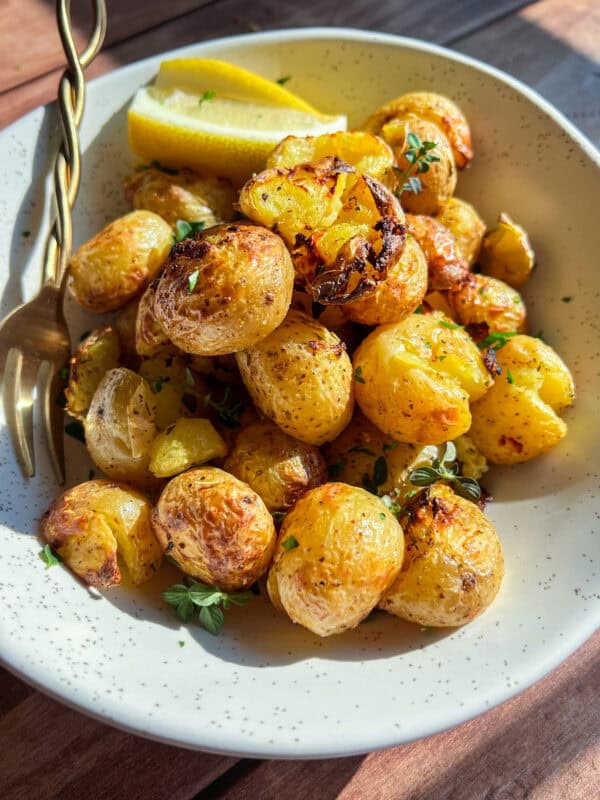 A white speckled bowl filled with roasted baby potatoes garnished with herbs and two lemon wedges, with a gold fork resting on the edge, on a wooden surface.