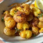 A bowl of crispy roasted baby potatoes garnished with fresh herbs, served with a lemon wedge and a fork on the side, all on a speckled ceramic plate.