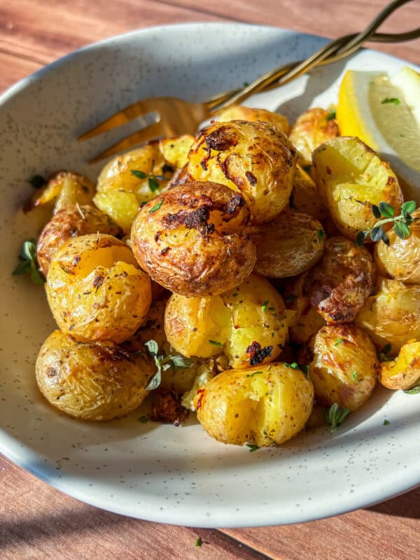 A bowl of crispy roasted baby potatoes garnished with fresh herbs, served with a lemon wedge and a fork on the side, all on a speckled ceramic plate.