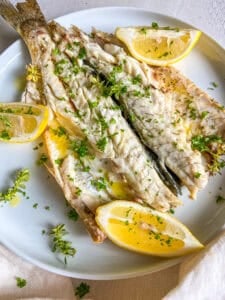 A plate with grilled fish garnished with chopped herbs and lemon wedges. The fish looks tender and seasoned, with olive oil drizzled on top. The lemons add a splash of color against the white plate.