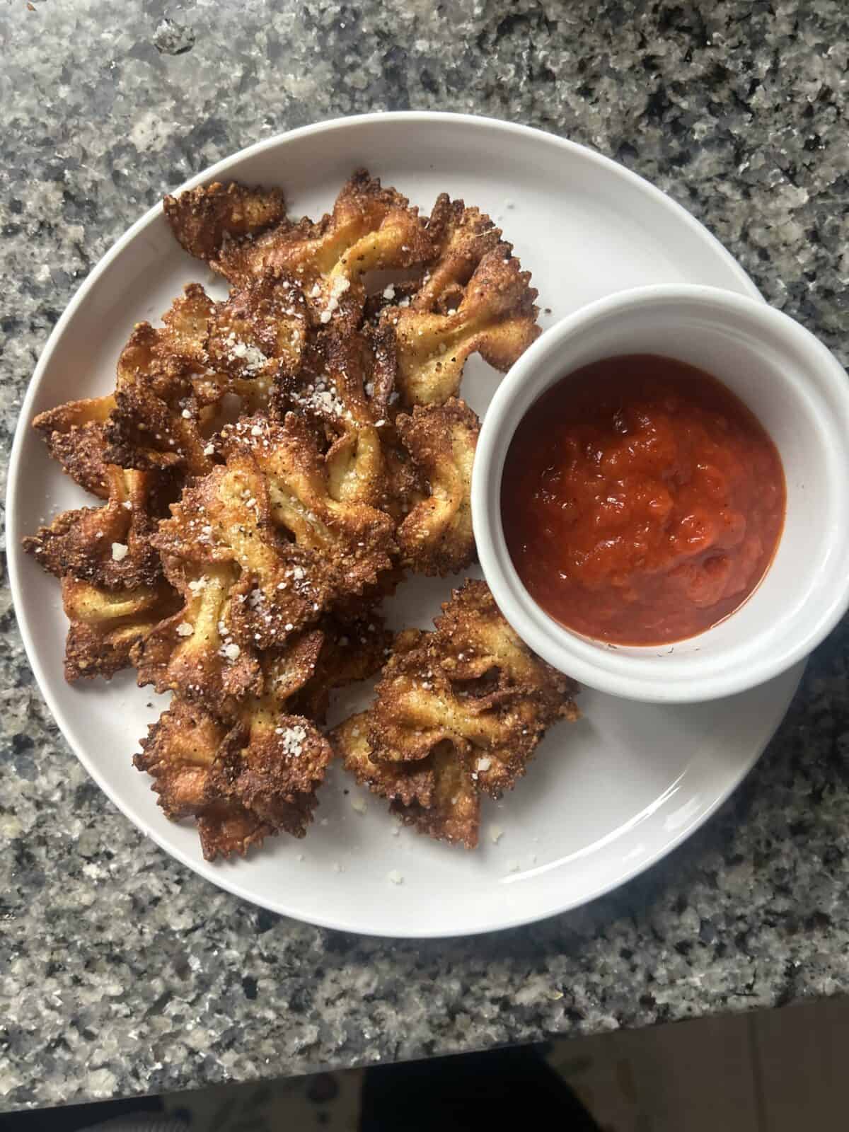 A white plate holds crispy, golden-brown Viral Pasta Chips sprinkled with grated cheese, served with a small ramekin of red marinara sauce on a gray, speckled countertop.