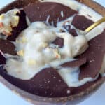A bowl filled with creamy banana ice cream, topped with large pieces of dark chocolate and sprinkled with chopped nuts, creates a Magic Shell Smoothie Bowl, served with a gold spoon resting inside.