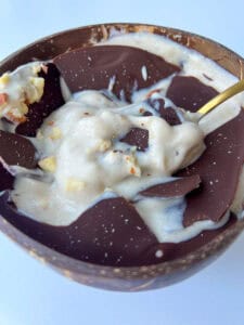 A bowl filled with creamy banana ice cream, topped with large pieces of dark chocolate and sprinkled with chopped nuts, creates a Magic Shell Smoothie Bowl, served with a gold spoon resting inside.