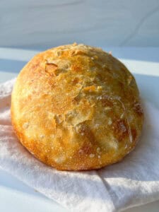 A round, golden-brown loaf of no knead bread sits on a folded white cloth, bathed in natural sunlight. The crust looks crispy with a slightly cracked surface.