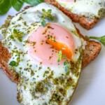 Two slices of toast topped with sunny-side-up eggs, green pesto, and sprinkled herbs, garnished with fresh basil leaves on a white plate. Try pairing with Air Fryer Hard Boiled Eggs for a delicious breakfast twist.