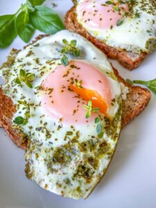 Two slices of toast topped with sunny-side-up eggs, green pesto, and sprinkled herbs, garnished with fresh basil leaves on a white plate. Try pairing with Air Fryer Hard Boiled Eggs for a delicious breakfast twist.