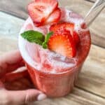 A hand holds a glass of iced strawberry drink topped with fresh strawberry slices and a mint sprig, with a clear straw, on a wooden table.