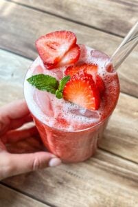 A hand holds a glass of iced strawberry drink topped with fresh strawberry slices and a mint sprig, with a clear straw, on a wooden table.
