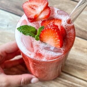 A hand holds a glass of iced strawberry drink topped with fresh strawberry slices and a mint sprig, with a clear straw, on a wooden table.