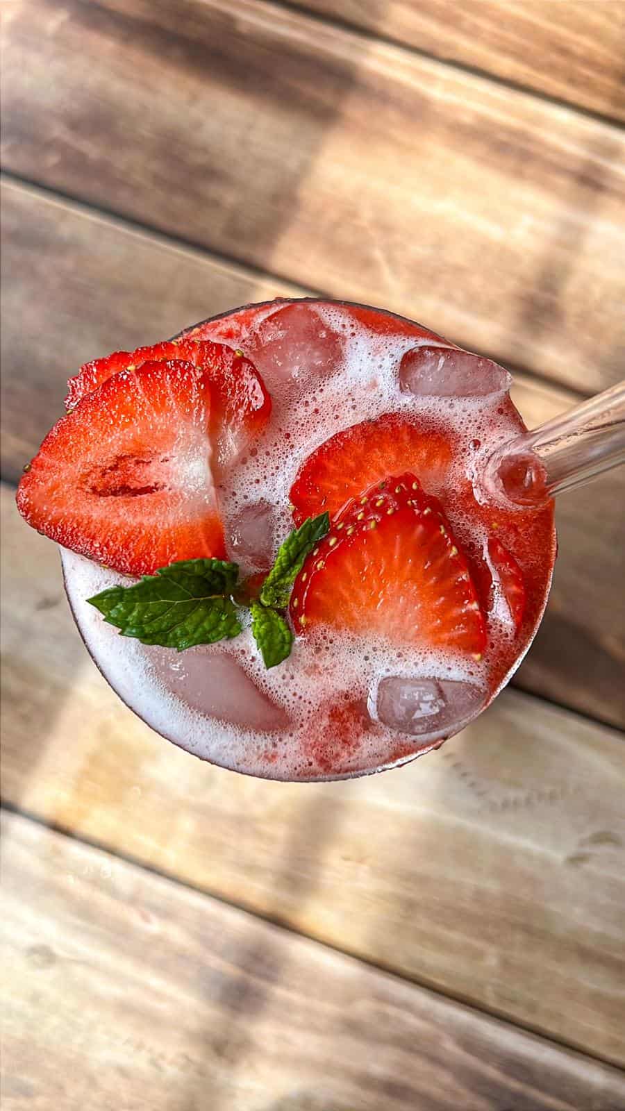 A refreshing drink with ice, garnished with fresh strawberry slices and a sprig of mint, served in a glass on a wooden surface.