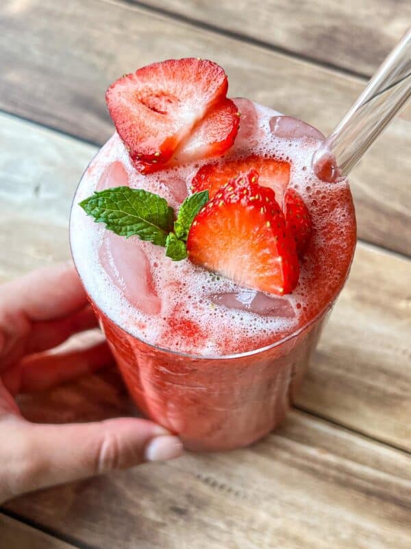 A hand holds a glass of iced strawberry drink topped with fresh strawberry slices and a mint sprig, with a clear straw, on a wooden table.
