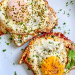 Two sunny-side-up eggs sprinkled with herbs and garnished with fresh green basil leaves on a white plate, served alongside flavorful Sun Dried Tomato Pesto Eggs for a burst of vibrant color and taste.