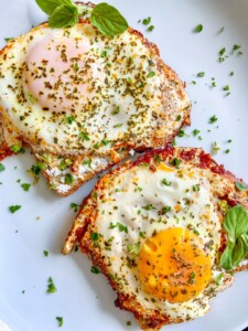 Two sunny-side-up eggs sprinkled with herbs and garnished with fresh green basil leaves on a white plate, served alongside flavorful Sun Dried Tomato Pesto Eggs for a burst of vibrant color and taste.