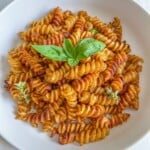 A white plate filled with rotini pasta in a red sauce, garnished with a sprig of fresh basil and a few small herb leaves, set on a light surface.