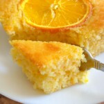 A close-up of a moist orange cake on a white plate, topped with a round slice of orange. A fork is holding a piece of the cake, showing its soft and fluffy texture.