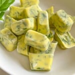 A white bowl filled with yellow cubes of frozen herb butter, with visible green herb pieces mixed in. A fresh basil leaf is on the side of the bowl.
