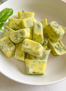 A white bowl filled with yellow cubes of frozen herb butter, with visible green herb pieces mixed in. A fresh basil leaf is on the side of the bowl.