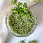 A glass jar filled with dried green herbs, topped with fresh flowering herb sprigs. The jar is placed on a light surface, with a white cloth and more fresh herbs nearby.