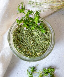 A glass jar filled with dried green herbs, topped with fresh flowering herb sprigs. The jar is placed on a light surface, with a white cloth and more fresh herbs nearby.
