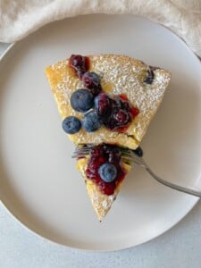A slice of cake topped with powdered sugar, blueberries, and berry sauce on a white plate. A fork is cutting into the cake.