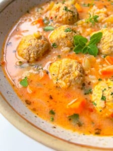 A close-up of a bowl of soup with meatballs, diced carrots, rice, herbs, and a creamy orange broth, garnished with fresh green herbs.