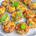 A white plate filled with baked stuffed mushrooms topped with melted cheese, chopped herbs, and garnished with fresh basil leaves.