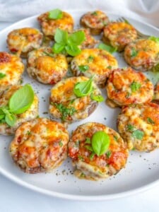 A white plate filled with stuffed mushrooms, topped with melted cheese, tomato sauce, herbs, and fresh basil leaves, arranged on a light background.