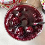 A close-up of a glass bowl filled with cherry preserves or compote, featuring whole cherries in a thick, glossy, deep red syrup. A spoon is partially submerged in the mixture. Jars in colorful fabric covers are nearby.