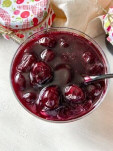 A close-up of a glass bowl filled with cherry preserves or compote, featuring whole cherries in a thick, glossy, deep red syrup. A spoon is partially submerged in the mixture. Jars in colorful fabric covers are nearby.