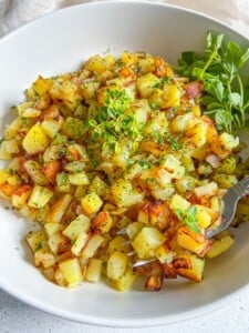 A white bowl filled with golden, diced roasted potatoes garnished with fresh green herbs, with a fork and a small bunch of leafy greens on the side.