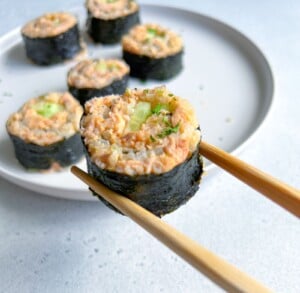 A pair of chopsticks holds a sushi roll with seaweed, rice, and a filling of salmon and cucumber. Several similar sushi pieces are arranged on a white plate in the background.