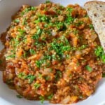 A white bowl filled with a chunky, colorful vegetable stew topped with chopped fresh herbs, served with a slice of multigrain bread on the side.