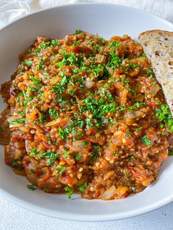 A white bowl filled with a chunky, colorful vegetable stew topped with chopped fresh herbs, served with a slice of multigrain bread on the side.