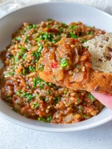 A close-up of a piece of bread topped with a chunky eggplant dip, garnished with chopped fresh herbs, held above a bowl filled with more of the same dip.
