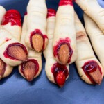 A close-up of spooky Witch Finger Cookies shaped like fingers, with almond slices for fingernails and red jam at the tips to resemble blood, arranged on a dark plate.