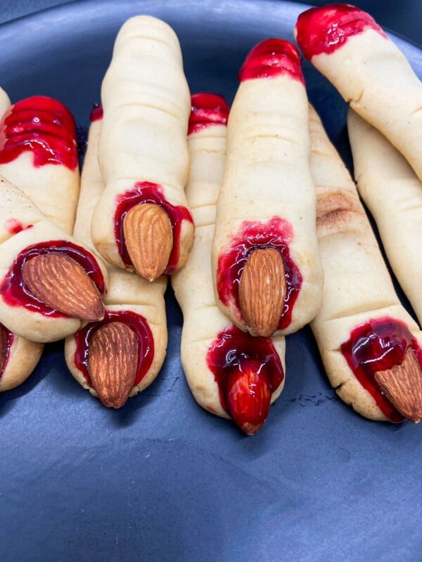 A close-up of spooky Witch Finger Cookies shaped like fingers, with almond slices for fingernails and red jam at the tips to resemble blood, arranged on a dark plate.