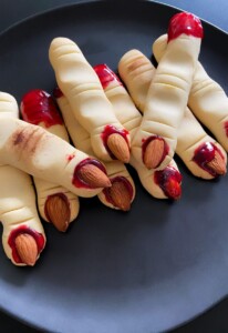 A black plate holds several Witch Finger Cookies shaped like severed fingers, with almond “nails” and red jam resembling blood at the tips and knuckles, creating a spooky Halloween treat.