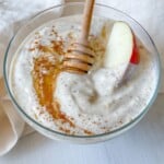 A glass bowl of creamy Yogurt Fruit Dip topped with honey, a sprinkle of cinnamon, and a slice of red apple. A honey dipper rests in the bowl, which sits on a white surface with a light cloth beside it.