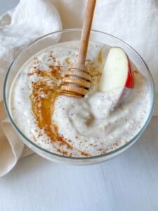 A glass bowl of creamy Yogurt Fruit Dip topped with honey, a sprinkle of cinnamon, and a slice of red apple. A honey dipper rests in the bowl, which sits on a white surface with a light cloth beside it.