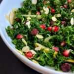 A close-up of a Kale Pomegranate Salad in a white bowl, topped with pomegranate seeds, dried cranberries, almond slices, and thin apple strips.