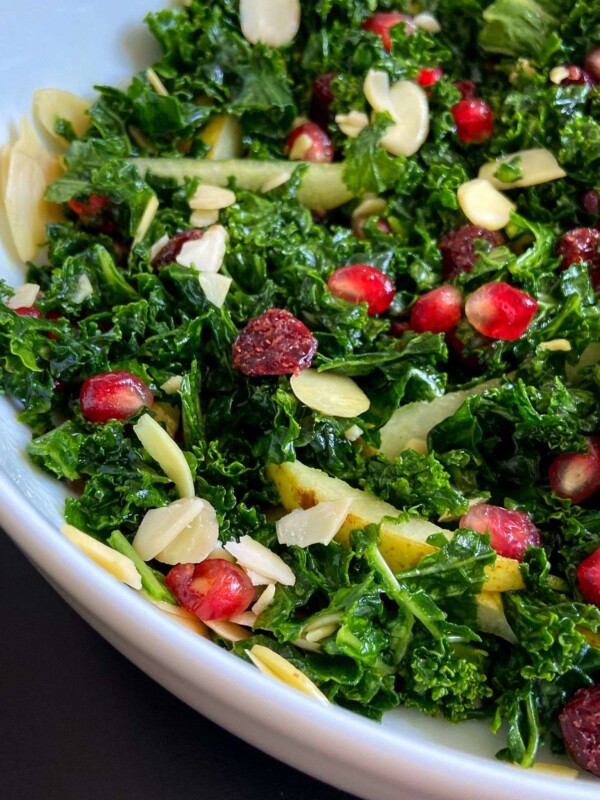 A close-up of a Kale Pomegranate Salad in a white bowl, topped with pomegranate seeds, dried cranberries, almond slices, and thin apple strips.